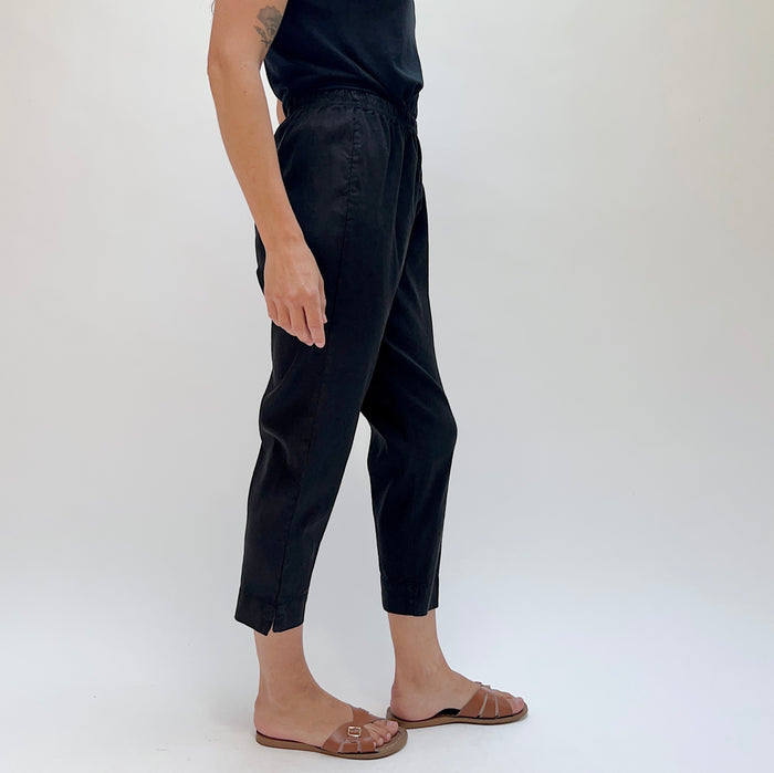Person wearing black cropped tapered pants and brown sandals with a white background, viewed from the side.