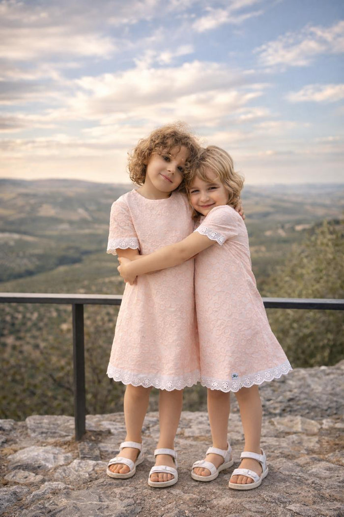 Two young girls with curly blonde hair share a hug while wearing matching Lola dresses in soft dusty pink. The dresses feature intricate floral lace embroidery throughout and delicate white scalloped eyelet lace trim on the short sleeves and hem. They are standing outdoors against a scenic mountain backdrop at sunset, wearing white sandals.