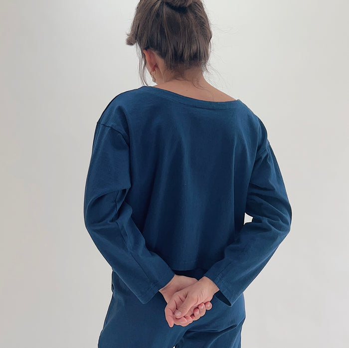 Back view of model wearing Pacific Cotton nova long sleeve button front shirt, showing wide back neckline, long sleeves, boxy cropped silhouette, banded hem, and deep teal color