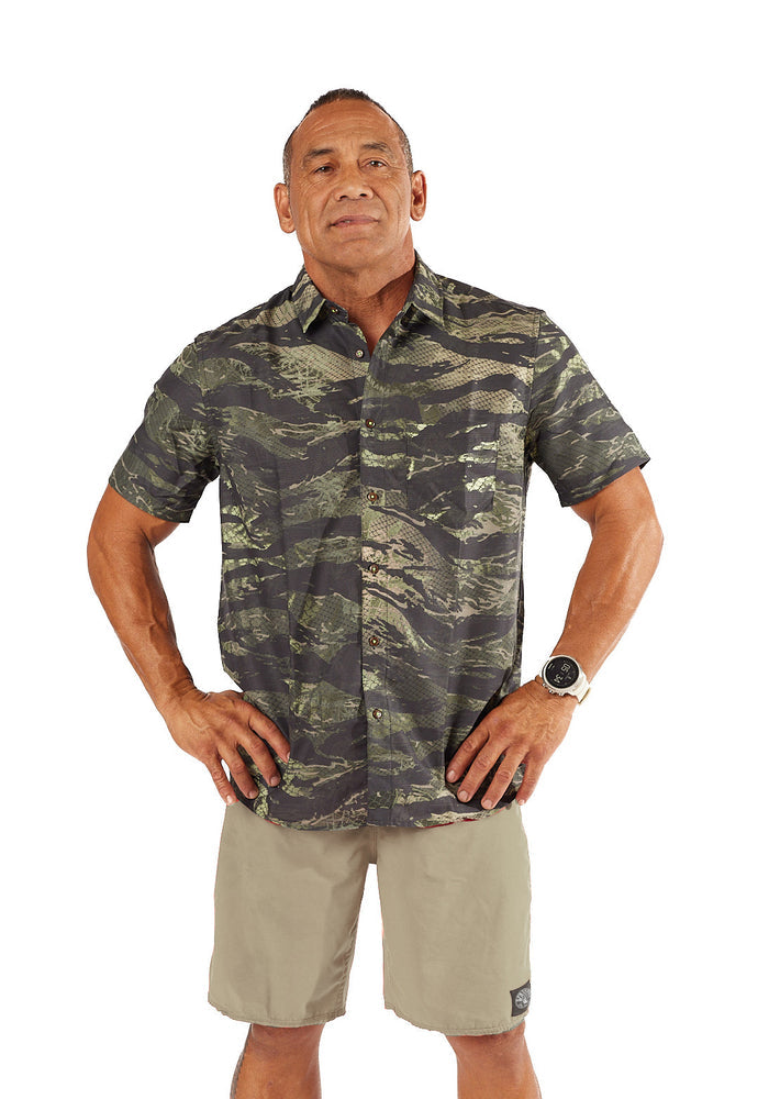Male model wearing the Waihana Men's Tropicam Aloha Shirt in Tropicam Green camouflage print, paired with khaki shorts, standing confidently with hands on hips against a white studio background.