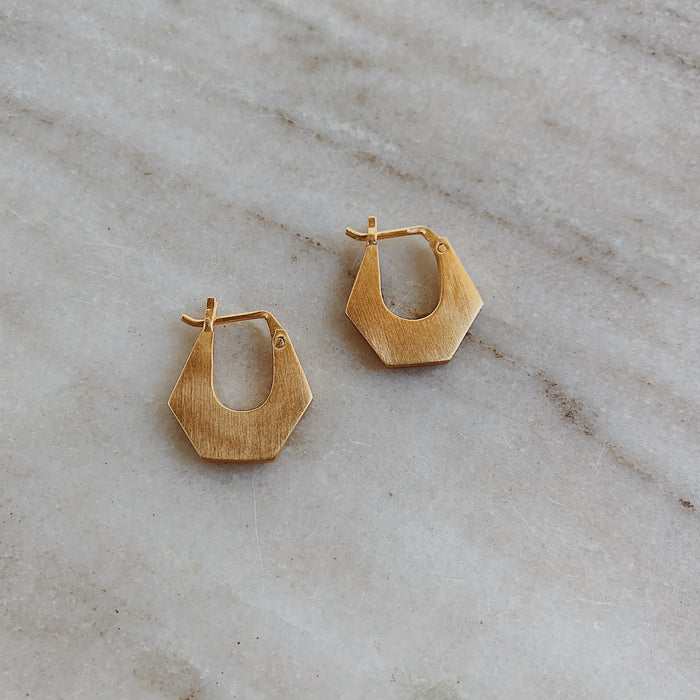 A pair of small chunky hexagonal gold hoop earrings on a marble surface.