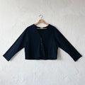 A black, long-sleeved crop cardigan with a button front, displayed on a hanger against a white textured wall.
