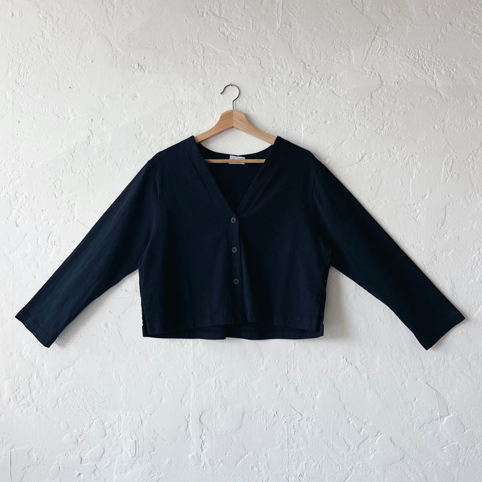 A black, long-sleeved crop cardigan with a button front, displayed on a hanger against a white textured wall.
