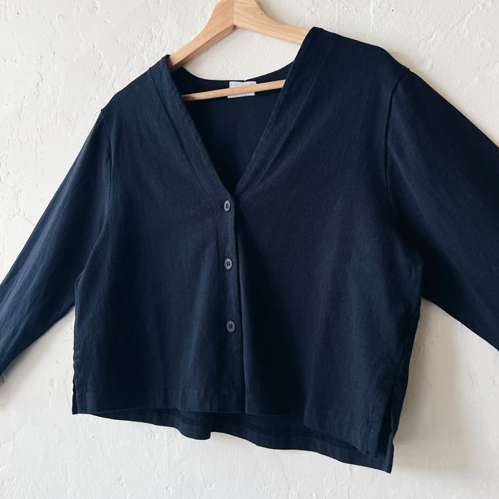 A black, long-sleeved crop cardigan with a button front, displayed on a hanger against a white textured wall.