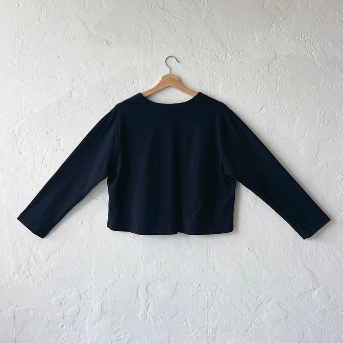 A black, long-sleeved crop cardigan with a button front, displayed on a hanger against a white textured wall, viewed from the back