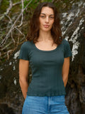 Female model wearing the Mollusk Surf Shop Pointelle Tee in Black Forest, a deep teal short-sleeve cotton tee with scoop neck and ruffled trim, styled with light blue jeans against a rocky outdoor backdrop