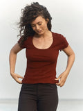 Female model wearing the Mollusk Surf Shop Pointelle Tee in Malbec, a deep red-brown short-sleeve cotton tee with scoop neck and ruffled trim, styled with black pants against a light grey background