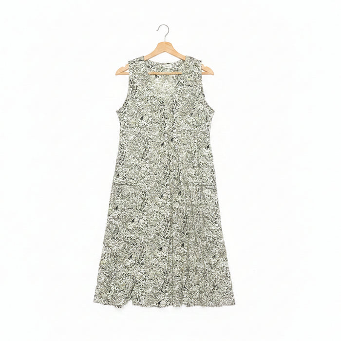 A sleeveless A-line dress with a dense green floral pattern on a white base, hanging on a wooden hanger on a dark concrete floor.