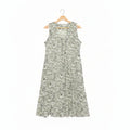 A sleeveless A-line dress with a dense green floral pattern on a white base, hanging on a wooden hanger on a dark concrete floor.