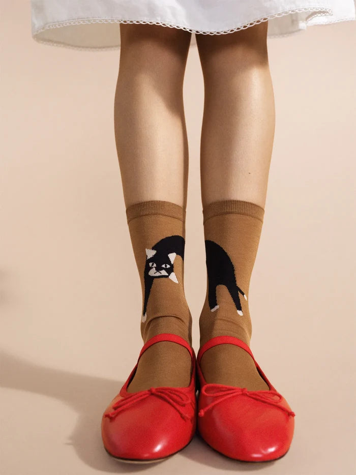 Pounce Crew socks in Camel — warm camel cotton crew with a black and white tuxedo cat motif, worn with red leather bow ballet flats under a white eyelet skirt on a cream background