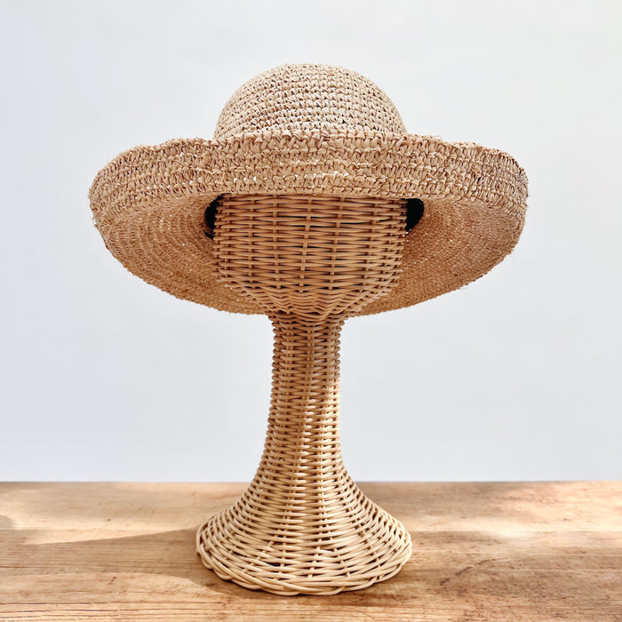 Natural raffia kettle brim hat with wide floppy brim and open block crochet weave, shown on a wicker hat stand — front view
