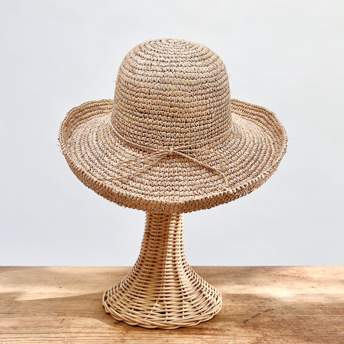 Natural raffia kettle brim hat with wide floppy brim and self-tie elastic band, shown on a wicker hat stand — side view