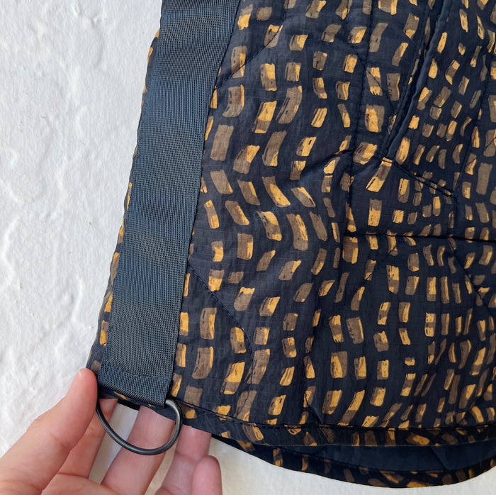 Black vest detail with gold geometric pattern held by a hand against a white background