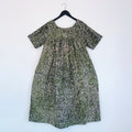 Full front view of the Sakina Dress in Green and Pink Speckle, hanging on a black hanger against a white wall, showing a round neckline, short sleeves, gathered yoke, voluminous A-line silhouette, and hand-printed green, pink, and white speckle pattern on 100% cotton Batiki fabric