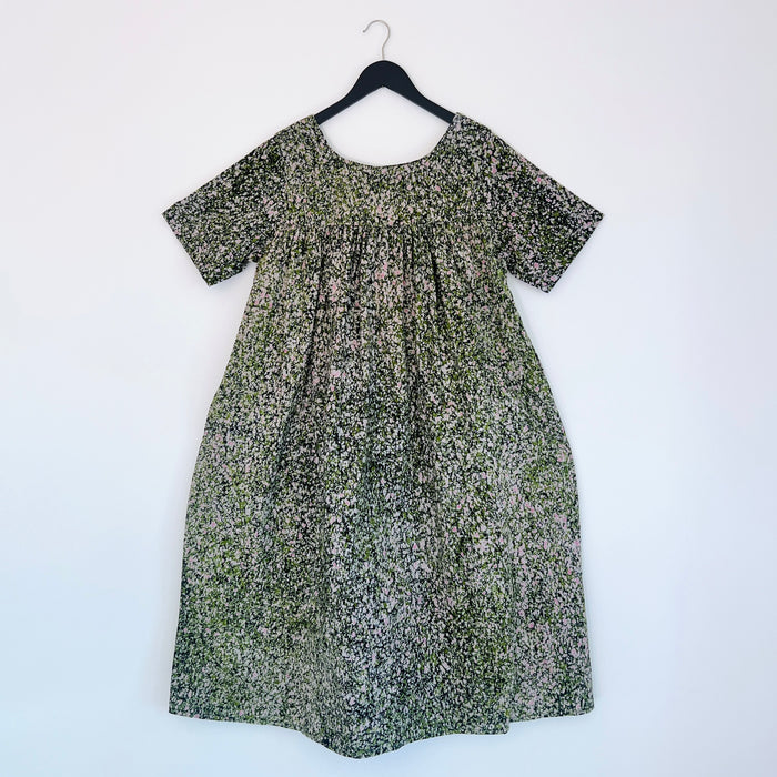 Full front view of the Sakina Dress in Green and Pink Speckle, hanging on a black hanger against a white wall, showing a round neckline, short sleeves, gathered yoke, voluminous A-line silhouette, and hand-printed green, pink, and white speckle pattern on 100% cotton Batiki fabric
