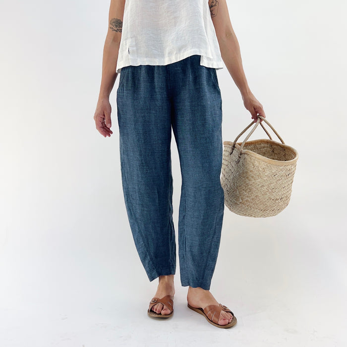 Person wearing blue melange linen pants and a white top and brown sandals, holding a woven basket on a white background.