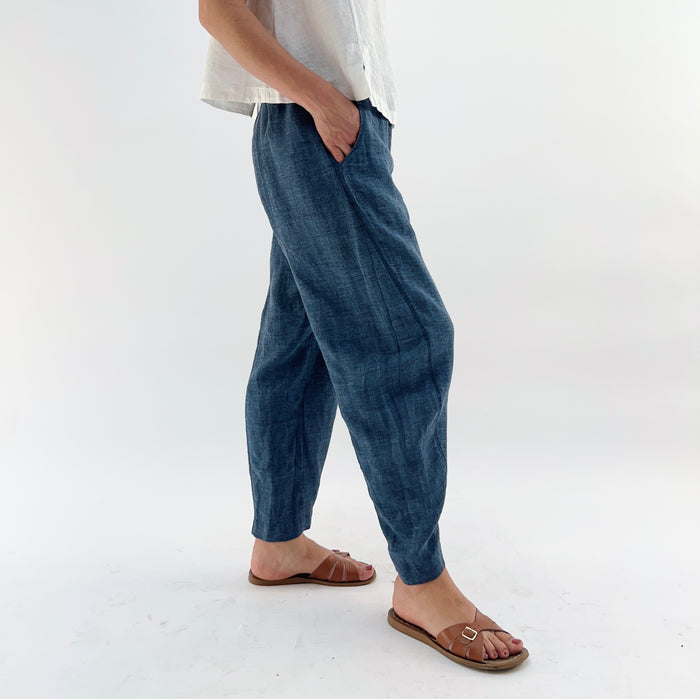 Person wearing blue melange linen pants and a white top and brown sandals, with their hands in their pockets on a white background.