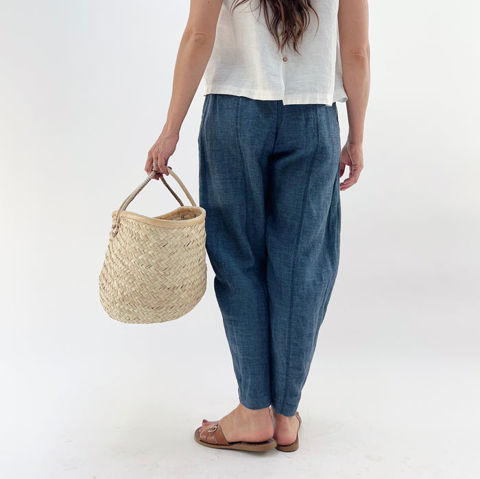 Person wearing blue melange linen pants and a white top and brown sandals, holding a woven basket on a white background as seen from behind.