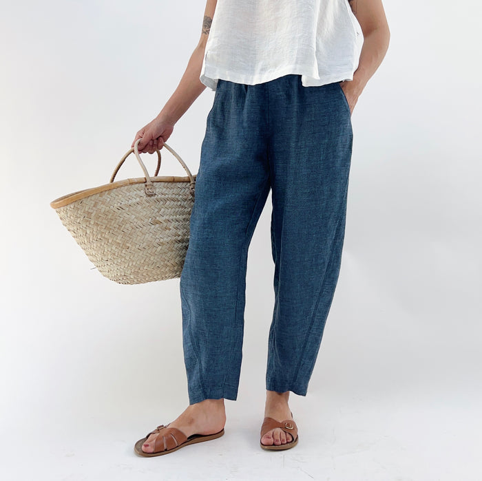 Person wearing blue melange linen pants and a white top and brown sandals, holding a woven basket on a white background.