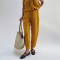 Three-quarter view of the Seamed Taper Pant in Tumeric by Yuvita, showing the tapered linen silhouette and seamed front leg in golden yellow, styled with a matching linen top, woven tote bag, and dark Mary Jane flats