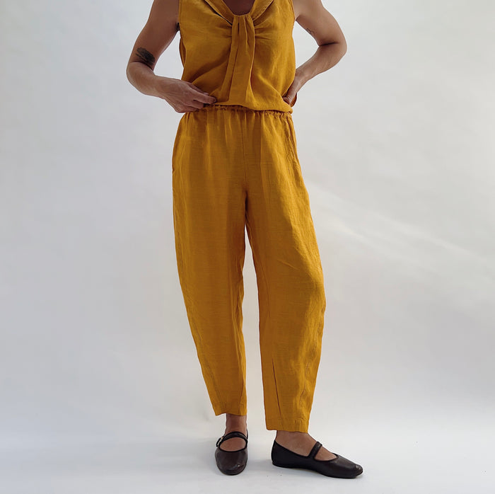 Front view of the Seamed Taper Pant in Tumeric by Yuvita, showing the elastic waistband, side pockets, and tapered ankle in golden yellow linen, styled with a matching tie-front linen top and dark Mary Jane flats