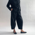 Three-quarter side view of model wearing Eleven Stitch black snap doreen pant, showing elastic waistband, wide balloon leg tapering to snap hem at ankle, side seam detailing, snap buttons at ankle, and black color, styled with matching black blossom collar shirt and black ballet flats