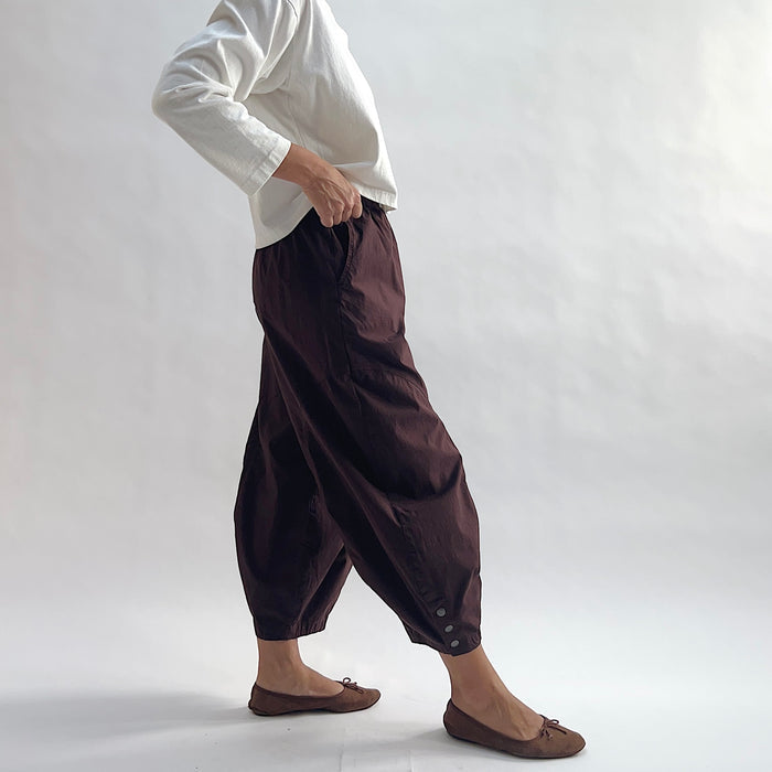 Side view of model wearing Eleven Stitch chicory snap doreen pant, showing elastic waistband, wide balloon leg tapering to snap hem at ankle, side seam pocket, and deep espresso brown color, styled with cream long sleeve top and taupe ballet flats