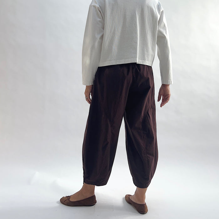 Back view of model wearing Eleven Stitch chicory snap doreen pant, showing elastic waistband, wide balloon leg tapering to snap hem at ankle, side seam detailing, and deep espresso brown color, styled with cream long sleeve top and taupe ballet flats