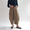 Close front view of model wearing Eleven Stitch winter olive snap front pant, showing snap front waistband, wide balloon leg, large side patch pockets, gathered elastic ankle hem, and warm olive tan color, styled with black button front top and black ballet flats