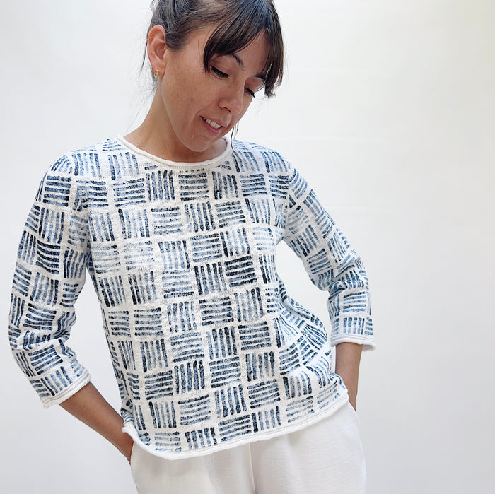 Woman wearing the Softly Stamped Pullover in White with hands in pockets, showing the relaxed fit, three-quarter sleeves, and navy stamp print, paired with white trousers