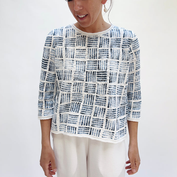 Smiling woman wearing the Softly Stamped Pullover in White, showing the full front view with round neckline, three-quarter sleeves, and navy brushstroke print, paired with white trousers
