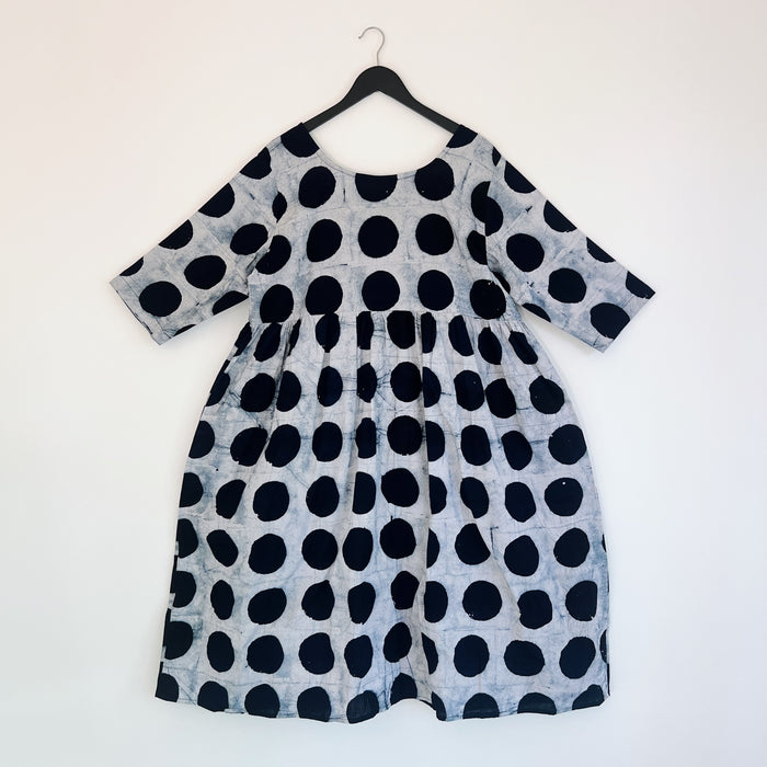 Full front view of the Sula Dress in Black Indigo Dots, hanging on a black hanger against a white wall, showing a round neckline, elbow-length sleeves, gathered waist seam, and oversized hand-printed black polka dots on grey-white 100% cotton Batiki fabric