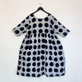 Full front view of the Sula Dress in Black Indigo Dots, hanging on a black hanger against a white wall, showing a round neckline, elbow-length sleeves, gathered waist seam, and oversized hand-printed black polka dots on grey-white 100% cotton Batiki fabric