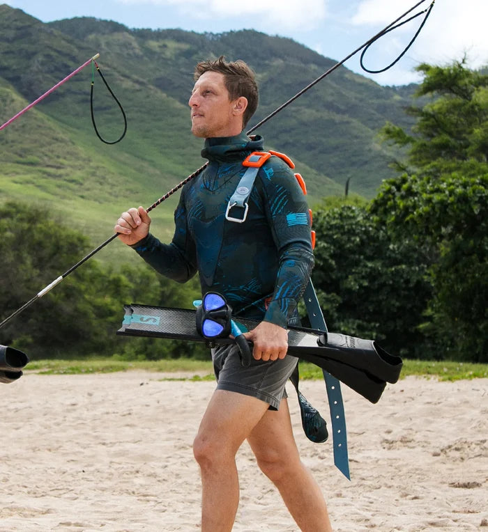 Male spearfisher walking on a sandy beach carrying carbon freediving fins, a blue dive mask, and a pole spear, wearing a dark patterned wetsuit top and an orange and teal weight belt, with lush green mountains behind him.