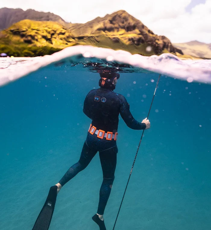 Male spearfisher wearing a dark wetsuit and Thunderstorm orange weight belt holds a pole spear in crystal-clear blue water, with lush Hawaiian mountains visible above the waterline in a dramatic split over-under perspective.