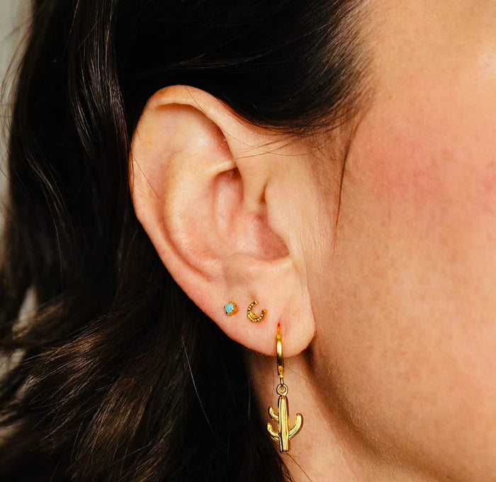 Close-up of an ear wearing gold hoop earrings with cactus charm and turquoise stone.