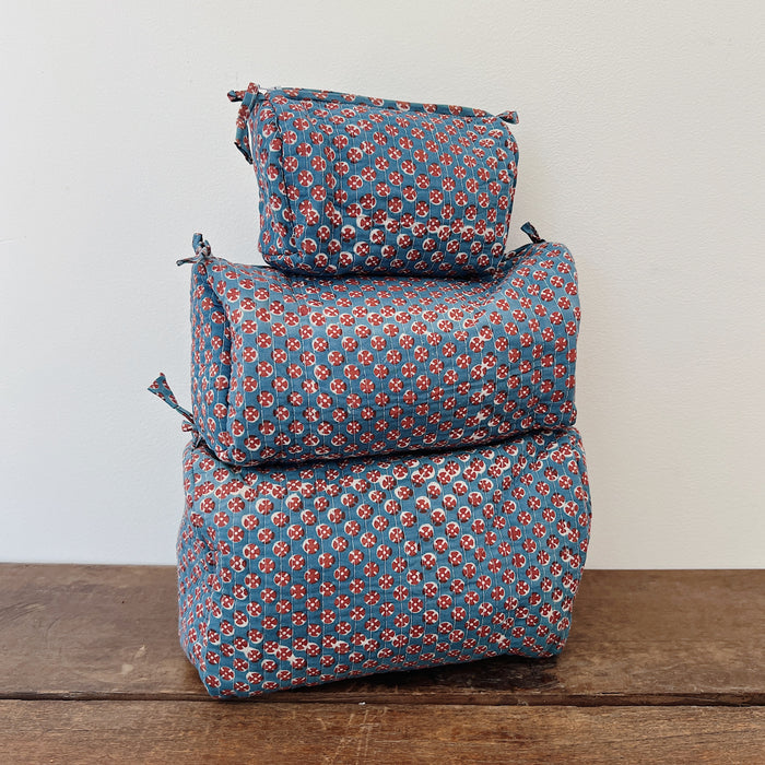 Three sizes of a quilted block-print toiletry bag stacked on a wooden surface — small, medium, and large — in a slate blue cotton with a red and cream circular block-print pattern