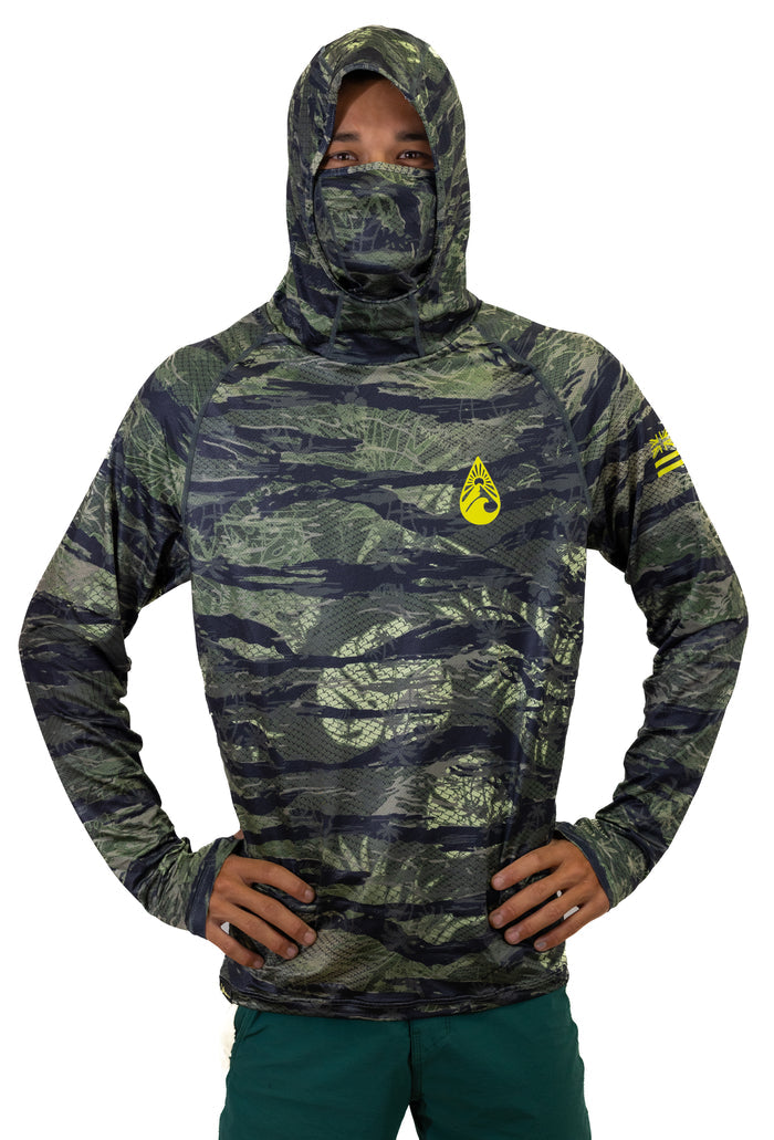 Male model wearing the Waihana Tropicam Sun Shirt in green and black camouflage pattern with attached hood and face mask pulled up, yellow Waihana brand logo on the chest, hands on hips against a white studio background.