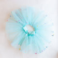 A turquoise tutu with multi-color confetti sprinkled on it, with an elastic waistband and a ribbon tie in the front.