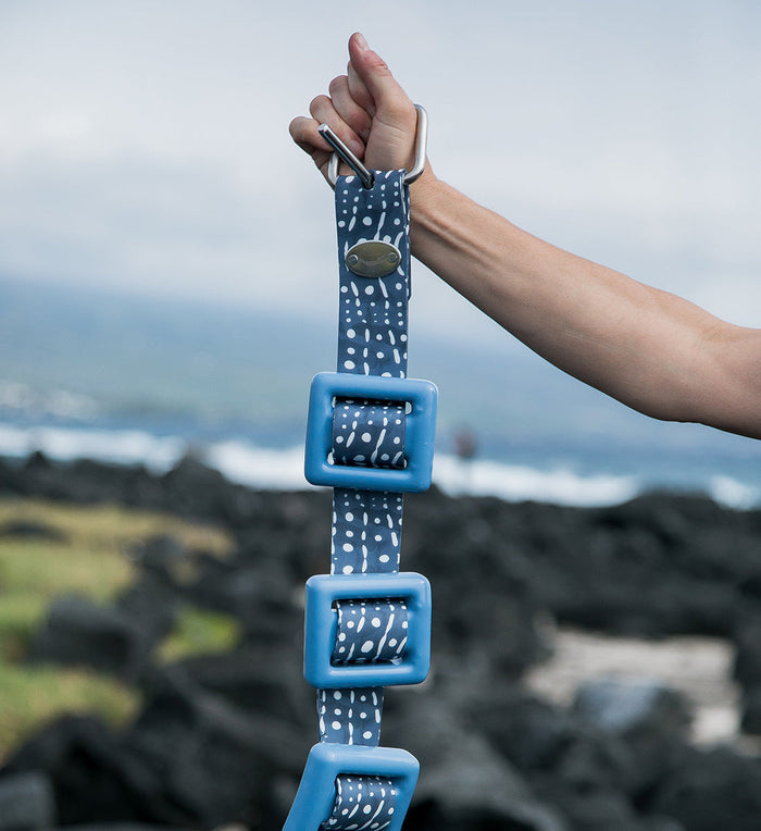 A hand holds up the Whale Shark Dive Belt featuring a navy blue and white spotted pattern with blue square weight retainers and a stainless steel buckle, with a rocky lava coastline and ocean visible in the background.