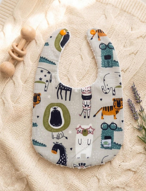 A flat lay of a light grey muslin baby bib featuring a whimsical "Zoo" print with colourful animals including a lion, tiger, zebra, and an elephant. The bib has orange snap closures and is displayed on a soft cream cable-knit blanket next to a wooden rattle and lavender sprigs.